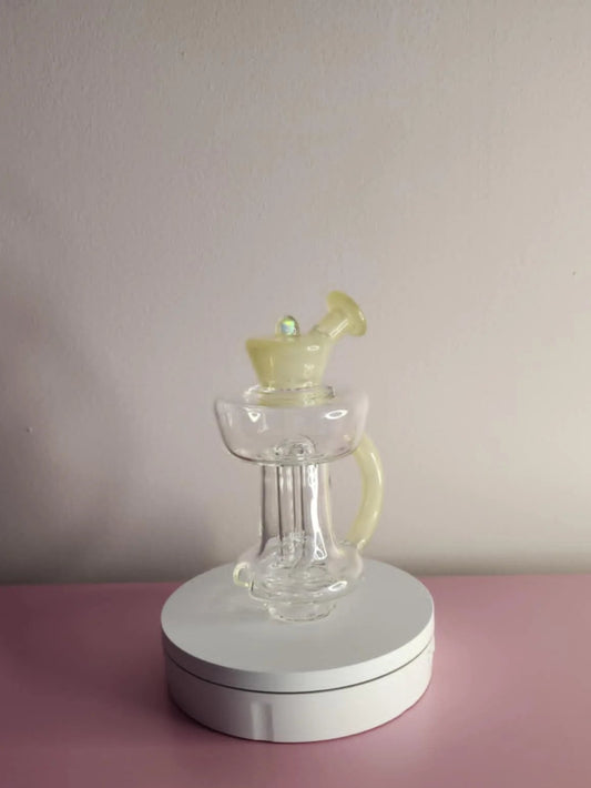 Clear glass bubbler with yellow handle on a white circular stand against a plain wall.